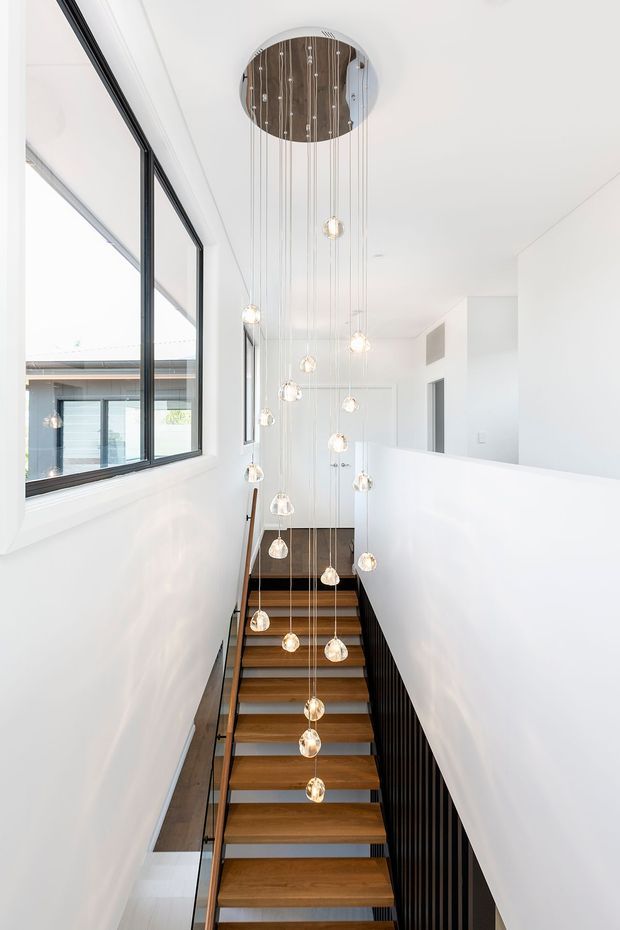 A bright modern stairway viewed from below, with open-riser timber treads, dark metal balustrades, white walls, and a dramatic cluster of globe pendant lights suspended from a circular ceiling feature. Black-framed glazing runs along the left side, bringing in natural light and revealing an adjacent exterior space.