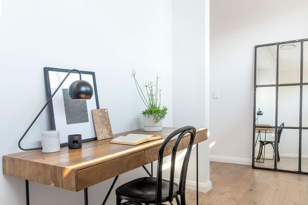 A bright, minimalist home office vignette with a substantial timber desk, black metal task lamp, framed artwork, small potted plant, and a black bentwood-style chair. A large industrial-style black-framed mirrored partition or door is visible to the right, reflecting part of the room. The space has white walls, timber flooring, and a clean contemporary aesthetic.