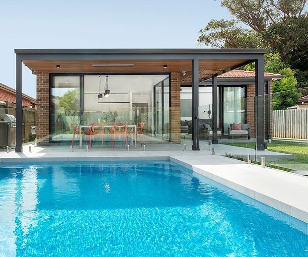 A contemporary single-storey glass-walled pool pavilion or extension overlooking a bright swimming pool. The structure has a flat dark metal roof and posts, warm timber soffit and ceiling lining, extensive black-framed glazing, and an open dining area with a long table inside. A covered outdoor lounge is visible to the right, with landscaped suburban surroundings and a tiled-roof house behind.