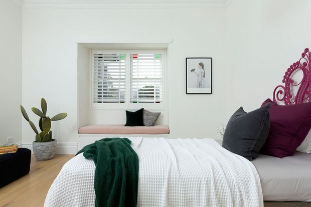 A bright, styled bedroom with a pale upholstered bed, white textured bedding, and deep green and burgundy accent cushions and throw. A pink window seat sits beneath plantation shutters, complemented by a potted cactus, framed artwork, ornate burgundy bedhead detailing, and decorative ceiling cornices.