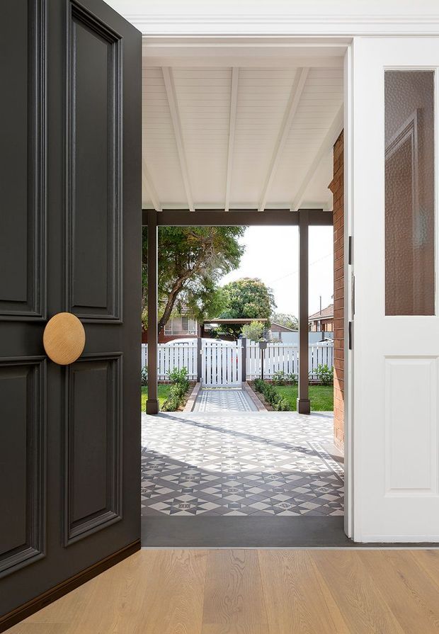 View from an interior entryway through an open front door to a covered porch and garden path. The entry features a light timber floor, a dramatic black paneled door with a round timber handle, a white glazed-panel door, and a patterned geometric tiled porch beneath a white-lined veranda roof. A white picket fence and landscaped front garden are visible beyond.