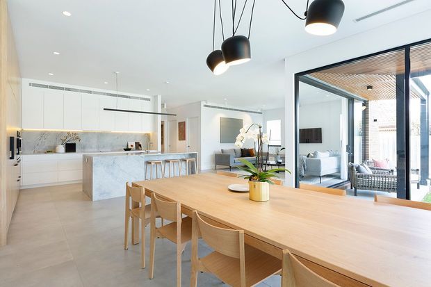 Bright open-plan interior featuring a contemporary white kitchen with a large pale stone island, a timber dining table with matching light-wood chairs, and a glazed opening to an outdoor area. Black sculptural pendant lights, pale flooring, concealed cabinetry, and a timber-lined exterior ceiling visible through the glazing create a clean, refined space.