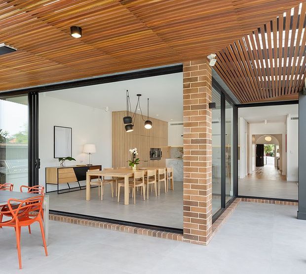 A bright contemporary indoor-outdoor dining and living area with extensive black-framed glazing and an open connection to a covered terrace. The space features a warm timber slatted ceiling and pergola, exposed face-brick columns and thresholds, pale concrete-look flooring, light timber kitchen cabinetry and dining furniture, and a long hallway extending through the home. Orange outdoor chairs provide a vivid accent.