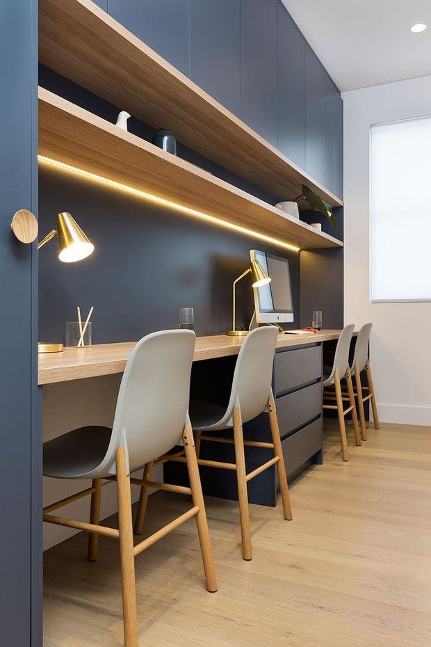 A compact, contemporary built-in study or work area with a long timber desk, dark blue-grey cabinetry and shelving, integrated warm LED shelf lighting, and multiple light upholstered timber-frame chairs. The joinery includes drawers and open display shelves with minimal decor, while a large window provides soft natural light. Pale timber flooring and clean lines create a refined, functional workspace.