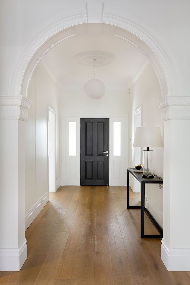 A bright, refined residential entry hall viewed toward a black paneled front door. The space features a broad white decorative archway with layered mouldings and pilasters, white walls and trim, a warm timber floor, sidelights beside the door, a simple spherical pendant, and a slim black console table with a large lamp.