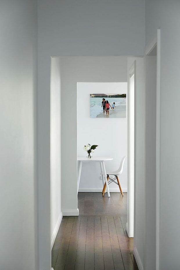 A narrow, bright white hallway with dark timber flooring leads through an open doorway into a minimalist dining or study nook. The adjoining room contains a small white table, a white molded chair with timber legs, a simple vase of flowers, and a framed beach photograph. White walls, trim, and door frames create a clean, airy interior.