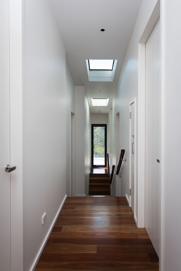 A narrow, bright residential hallway with white walls and ceiling, multiple white doors, dark timber flooring, recessed downlights, and two skylights bringing daylight into the corridor. The hall leads down a short flight of timber stairs toward a glazed exterior door, with a simple timber handrail visible on the right.