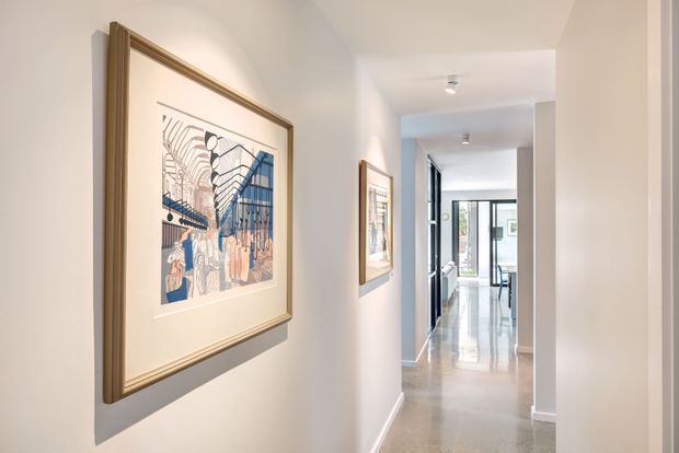 A bright, narrow residential hallway with white walls, recessed ceiling lighting, framed artwork, and polished light-coloured flooring. The corridor leads toward a glazed living or dining area at the rear, creating a strong sense of depth and natural light.
