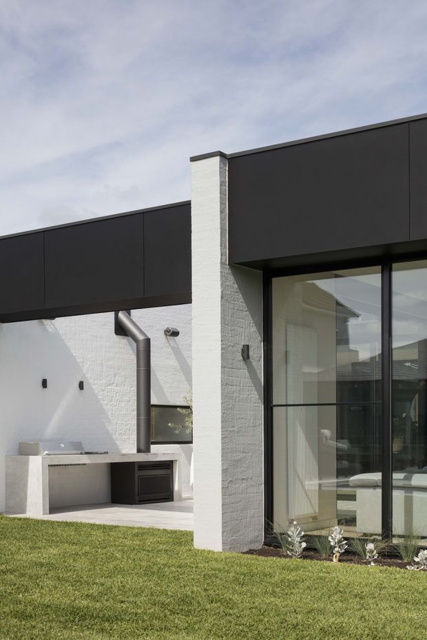 Exterior view of a contemporary single-storey residence with a crisp, rectilinear composition. Dark charcoal flat roof volumes contrast with white textured masonry walls, while a large black-framed glazed opening reveals a light-filled interior. A covered outdoor cooking area with a built-in barbecue and metal flue sits to the left, opening onto a neatly landscaped lawn.