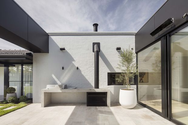 A bright contemporary courtyard or outdoor entertaining area centered on a white rendered masonry wall with a built-in outdoor fireplace, tall black flue, and pale stone or concrete bench. Black-framed glazed doors and windows open onto the courtyard from the surrounding home. The space has light paving, minimalist detailing, and a large white planter with a small tree.