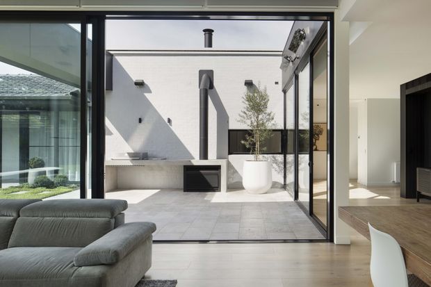Contemporary interior looking through expansive black-framed glazing onto a private paved courtyard. A white rendered courtyard wall features an outdoor fireplace or chimney structure, with a large white planter and small tree. The foreground contains a grey sectional sofa, pale timber flooring, and a partial white dining chair, while a dark built-in feature wall is visible to the right. The composition is clean, minimal, and strongly connected to the courtyard through full-height sliding doors.