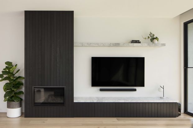 Contemporary living room media wall with a dark vertical-slatted feature surround, integrated fireplace, wall-mounted television and long low storage cabinet. Pale marble-look shelving and benchtop contrast with the dark joinery, while a potted plant and black-framed glazing add softness and natural light. Light timber flooring and warm neutral walls create a clean, refined interior.