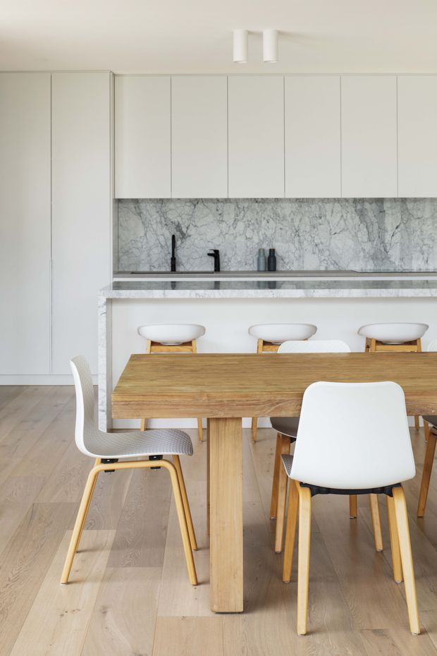 A bright, minimalist open-plan kitchen and dining area. Floor-to-ceiling white cabinetry surrounds a marble-look stone backsplash and matching island or peninsula benchtop, with a black tap and small appliances. A natural timber dining table is paired with white molded chairs featuring light timber legs. Pale timber flooring and a white ceiling create a clean, airy Scandinavian-inspired interior.