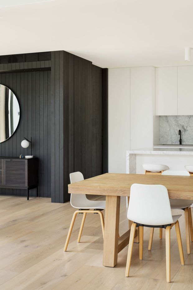 A bright, refined open-plan dining and kitchen interior. A substantial light timber dining table with white molded chairs occupies the foreground. Behind it, a white kitchen with marble-look stone benchtops and splashback is paired with a dramatic dark vertical timber-panel feature wall, integrated cabinetry, a black tap, and a round mirror above a dark timber console. Pale timber flooring and a white ceiling create a clean, balanced contrast.