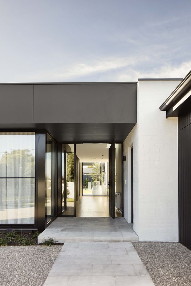 Contemporary residential entrance viewed from the front, with a broad dark cantilevered roof or soffit over a recessed glazed entry. The facade combines white painted brickwork, dark metal cladding and extensive black-framed glazing. A pale concrete path and steps lead through a minimalist gravel and planted forecourt into a bright interior corridor.