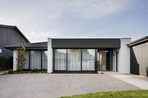 Street-facing view of a contemporary single-storey residence with a low, rectilinear form. The facade combines dark grey rendered walls, white rendered sections, extensive black-framed floor-to-ceiling glazing, a covered entry, and a large paved driveway. A pitched tiled roof is visible over the older adjoining section, while the main volume has a flat roofline.
