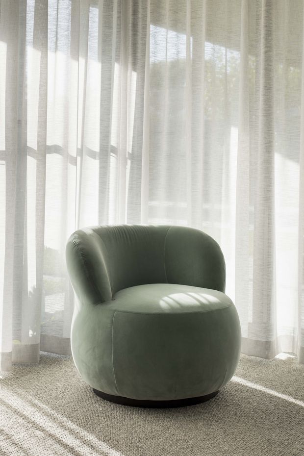 A softly lit interior vignette featuring a rounded, sculptural olive-green upholstered armchair positioned in front of full-height sheer white curtains. Sunlight filters through the curtains, casting linear shadows across the chair and neutral textured carpet. The composition is minimal and serene, with no other furnishings visible.