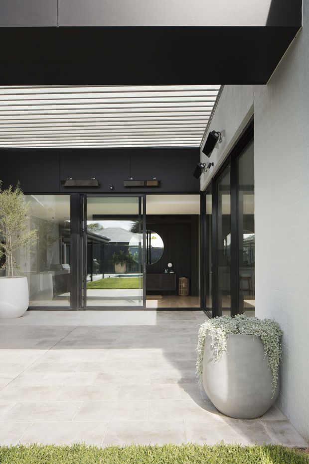 A modern residential courtyard or covered patio viewed from outdoors. Large pale stone or concrete-look paving extends toward black-framed glazed doors and windows. A white rendered wall, dark charcoal feature wall, and a broad translucent slatted pergola roof create a sheltered contemporary outdoor room. Large minimalist planters flank the glazing, with an interior living area visible beyond the open doors. The composition is bright, clean-lined, and strongly connected to the garden.