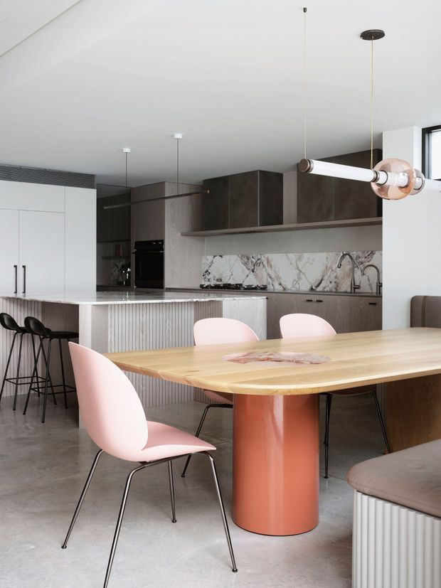 Contemporary open-plan kitchen and dining area with a large light timber dining table, curved terracotta pedestal base, pale pink dining chairs, and a kitchen island with ribbed detailing. The kitchen features dark timber cabinetry, white cabinetry, a marble-look stone splashback, integrated appliances, concrete-look flooring, and a sculptural linear pendant light.