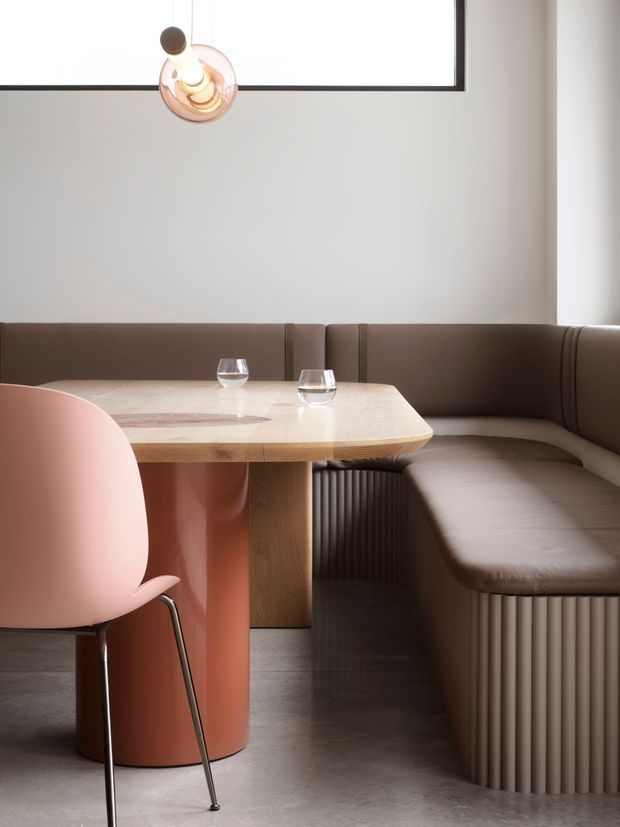A refined contemporary dining nook with a curved built-in brown banquette wrapping around a light wood and stone-topped table. A blush pink dining chair, cylindrical terracotta-toned table pedestal, ribbed banquette bases, polished concrete floor, pale plaster walls, and a sculptural pendant light create a warm, minimalist interior.