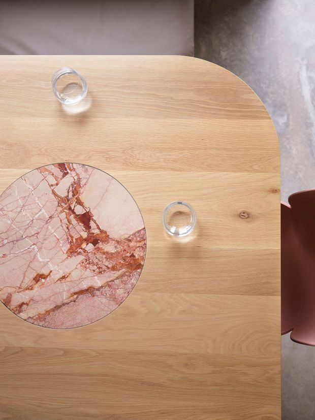 Top-down view of a light natural timber dining table with rounded corners, featuring a circular pink and burgundy veined stone inset. Two clear drinking glasses sit on the table, with partial muted pink dining chairs visible at the edges and a neutral floor and wall background.