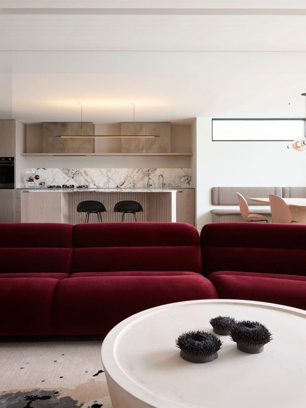 A refined open-plan living, dining and kitchen interior. A deep burgundy modular sofa dominates the foreground, facing a large rounded white coffee table with sculptural dark objects. Behind it is a compact kitchen with light timber cabinetry, a marble-look stone splashback and island with black bar stools. The adjacent dining nook has a built-in upholstered bench, rounded blush dining chairs and a linear horizontal window. The space has a low, smooth white ceiling with concealed lighting and a restrained contemporary palette.