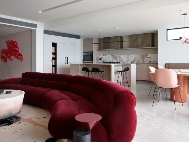 Open-plan contemporary living, dining and kitchen interior with a large deep burgundy curved sectional sofa in the foreground. The kitchen features pale wood cabinetry, a stone-look island benchtop, integrated appliances and dark bar stools, while a dining area with blush pink chairs sits beside the kitchen. White walls, pale flooring, linear ceiling detailing and a small abstract red wall artwork create a refined modern setting.