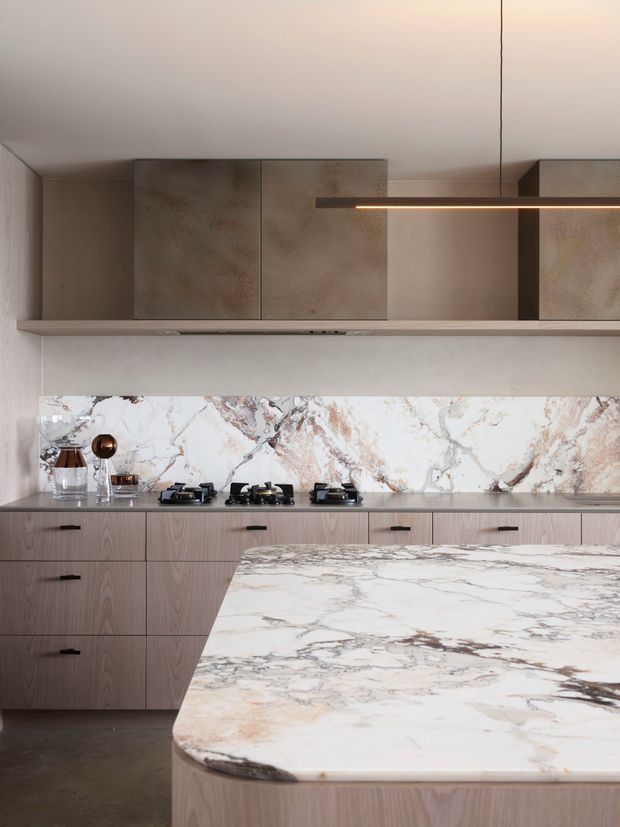 Contemporary kitchen with a large waterfall-edge island, pale wood-grain cabinetry, a dramatic veined stone benchtop and splashback, integrated upper storage, and a slim linear pendant light. The palette is warm and muted with refined natural materials and metal accents.