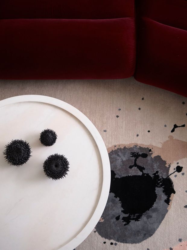 Top-down detail of a refined interior vignette featuring a deep burgundy upholstered sofa, a pale round tray or tabletop, three black spiky decorative objects, and a neutral abstract patterned rug with dark charcoal and black forms.