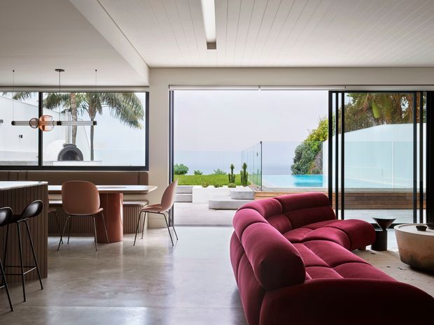 Open-plan contemporary living and dining space with a sculptural deep burgundy sectional sofa, compact dining area, and kitchen island seating. Full-height black-framed glazing opens toward a pool and ocean-facing outdoor terrace. The room features polished concrete flooring, white painted timber ceiling boards, and minimalist built-in joinery.