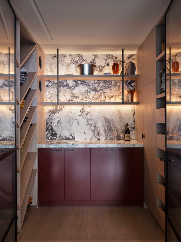 A compact, highly finished wine room or bar with floor-to-ceiling timber shelving, integrated bottle storage, dark burgundy cabinetry, a dramatic veined stone splashback and benchtop, and warm integrated shelf lighting. Decorative glassware and bottles are displayed on open shelves, creating an intimate, luxurious atmosphere.