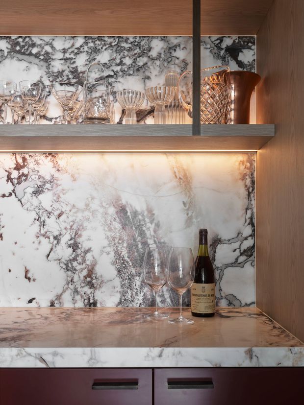 Close-up of a luxurious built-in bar and glassware display. Veined white marble or stone forms the backsplash, shelf surfaces and counter, framed by warm timber cabinetry and dark burgundy lower drawers. Integrated warm LED lighting illuminates crystal glasses, decanters, a copper-toned pitcher and wine bottle.