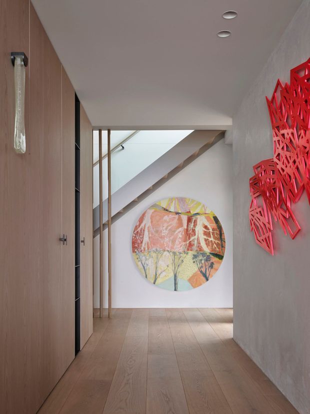 A contemporary residential hallway with wide light timber floorboards, full-height timber cabinetry along one side, white plaster walls, and recessed ceiling lights. A staircase rises at the far end behind slender vertical timber screens, while colourful circular artwork and vivid red geometric wall art add strong decorative accents.