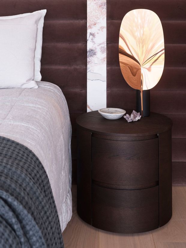 Close-up bedroom scene featuring a partially visible bed with white bedding and a dark textured throw, a deep brown upholstered wall or headboard, and a rounded dark timber bedside table. The table holds a sculptural oval lamp with a warm abstract shade, a small ceramic bowl, and decorative crystals. A narrow marble-look vertical wall panel adds contrast.