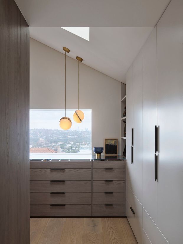 A compact, refined dressing and storage area with floor-to-ceiling white built-in wardrobes on the right, a dark timber-toned bank of drawers beneath a large window, and open shelving beside it. Two warm pendant lights hang over the joinery. Pale timber flooring, neutral walls, and a distant urban hillside view create a bright, minimalist interior.