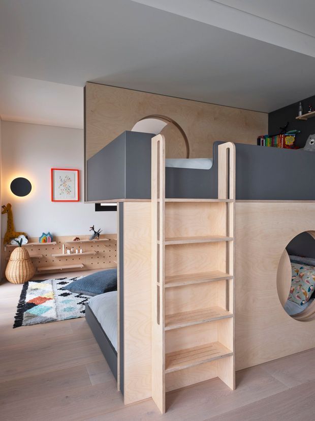 A playful children's bedroom with a custom plywood bunk-bed structure featuring integrated ladder shelving, rounded arch openings, and dark grey panels. The room includes a low pegboard storage unit, colourful toys and books, a patterned rug, and soft neutral finishes under a sloped ceiling.