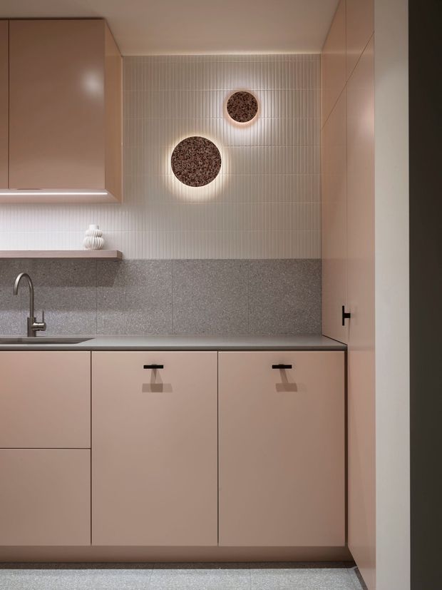 Compact, warmly lit contemporary kitchen or kitchenette with muted blush-beige cabinetry, a dark speckled stone benchtop and matching low backsplash, and vertically ribbed pale wall tiles. Integrated under-cabinet lighting highlights the work area, with a slim gooseneck tap and sink at left. Two circular textured wall lights or decorative features add a soft focal point.