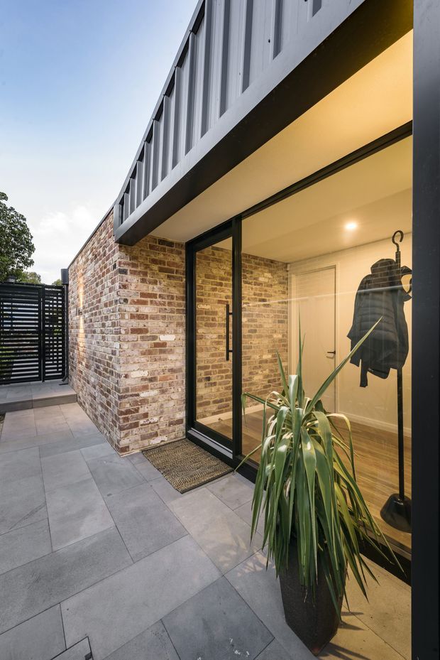 A contemporary residential entry courtyard viewed from outdoors, with a paved grey-tile walkway beside a warm-toned exposed brick wall and full-height black-framed glazing. The glazed entry reveals a warmly lit interior with timber flooring, white walls, and a coat rack. A tall potted plant sits near the doorway, while a dark slatted gate closes the courtyard at the rear. The upper storey projects above the entry with dark cladding and vertical window elements.