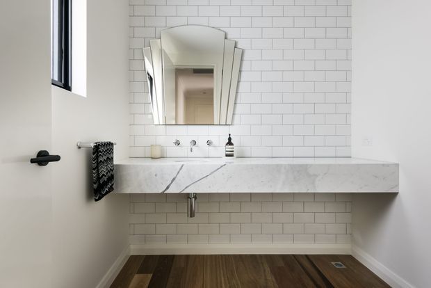 A compact contemporary bathroom featuring a wall-mounted pale stone vanity with integrated basin, white subway-tile walls, a geometric framed mirror, exposed plumbing, and dark timber flooring. The space is bright and minimal with black hardware and a small towel rail.