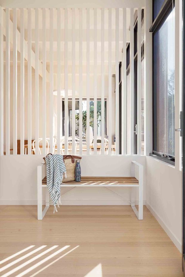 A bright, narrow interior passage or entry area with pale timber flooring, white walls, and a built-in timber-slat screen separating it from a light-filled living space beyond. A simple built-in bench with white supports and a timber seat sits beneath large dark-framed windows, with woven baskets and a striped throw adding subtle texture. Strong sunlight creates linear shadows across the floor, giving the space an airy, calm, coastal feel.