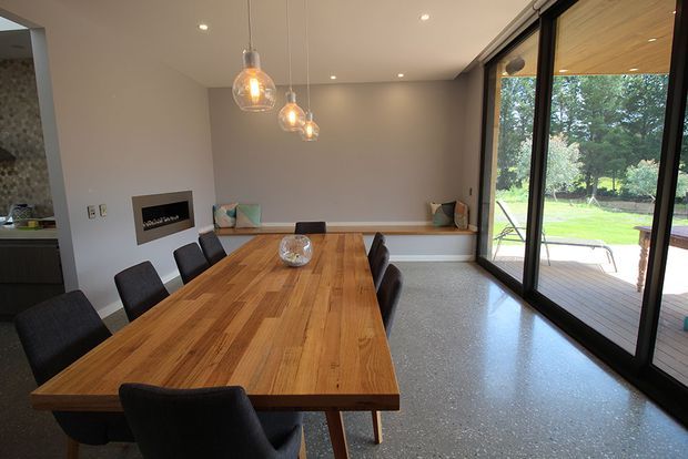 A contemporary dining room with a long solid timber dining table and dark upholstered chairs. The room features a built-in bench seat with cushions, three pendant lights, polished concrete or terrazzo-style flooring, neutral walls, and expansive black-framed sliding glass doors opening to a timber deck and garden.