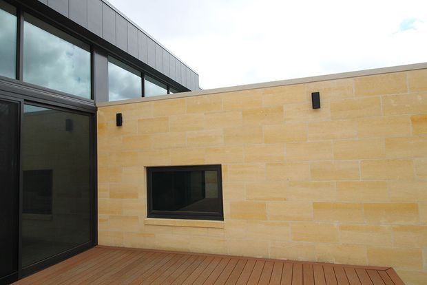 Modern courtyard-style exterior featuring a warm sandstone masonry wall with a small dark-framed window, dark metal-framed glazing and upper cladding, and a timber-toned deck. The composition highlights the contrast between the pale stone wall and contemporary charcoal glazing.