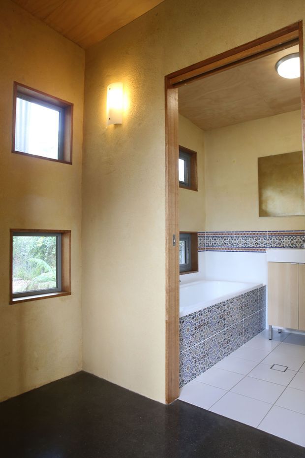 A warm-toned interior view of a small entry or hallway opening directly into a bathroom. The hallway has textured ochre plaster walls, a timber-lined ceiling, dark flooring, timber-framed windows, and a wall sconce. The bathroom features a white bathtub with patterned blue, white and terracotta ceramic tile surrounds, white floor tiles, timber cabinetry, and a large wall mirror.