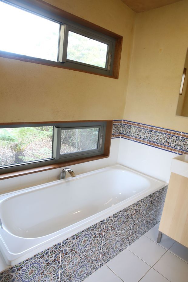 Bright bathroom featuring a built-in white bathtub beneath two horizontal windows. The tub surround and backsplash are clad in intricate blue, white, and terracotta patterned tiles, with warm timber window reveals and a light tiled floor. A partial vanity is visible at the right edge.