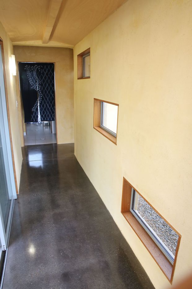 A narrow interior hallway with warm ochre plaster walls, a timber-lined ceiling, polished dark concrete flooring, and several small horizontal timber-framed windows. The corridor leads toward a tiled room screened by a dark patterned curtain.