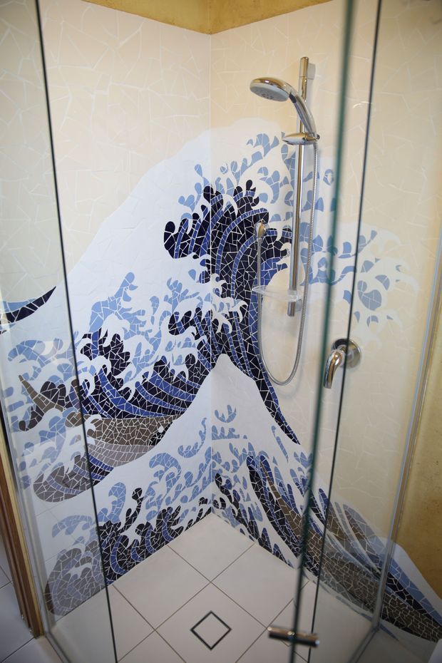 A compact enclosed shower with clear glass doors, chrome shower hardware, and highly decorative mosaic wall tiling depicting stylised dark blue and pale blue aquatic or floral forms on a cream background. The shower floor is finished with large light tiles and a small contrasting diamond inset drain detail.