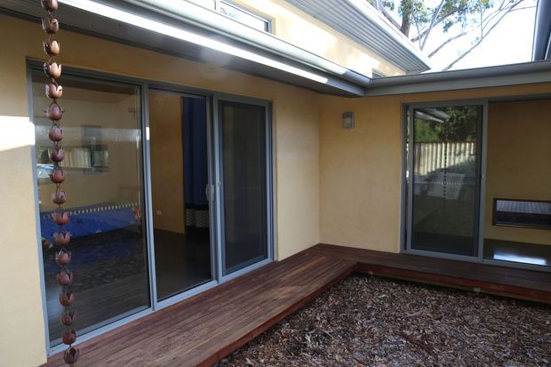 A small enclosed courtyard viewed from outdoors, surrounded by warm ochre rendered walls with dark aluminium-framed sliding glass doors and windows. Timber decking lines the perimeter around a central bed of bark mulch, with a hanging decorative feature at left and an integrated low window seat or bench along the right wall. The courtyard is sheltered beneath overhanging roof forms and clerestory-like glazing.