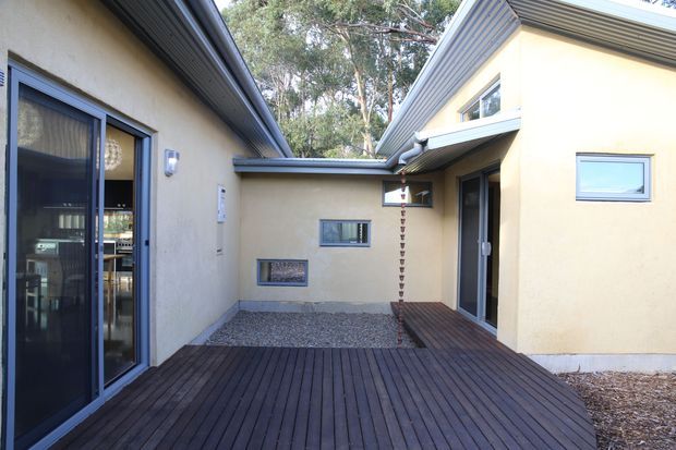 Exterior view of a compact courtyard formed by connected rendered buildings. A dark timber deck spans the foreground, with dark-framed glazed doors, small horizontal windows, corrugated metal roof edges and a narrow gravel area. Mature trees are visible beyond the courtyard.