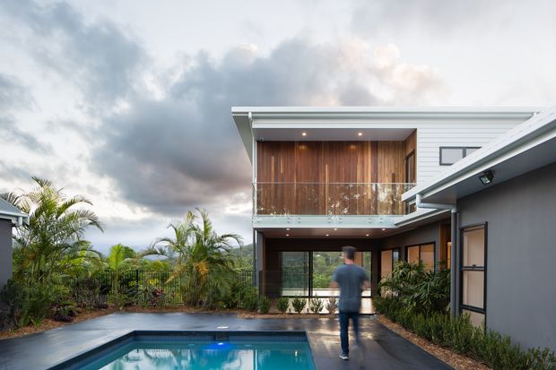 Contemporary two-storey residential home surrounding a compact swimming pool and landscaped courtyard. The house features crisp white horizontal cladding, dark grey rendered walls, broad roof overhangs, extensive glazing, and a warm timber-lined upper balcony facade with a glass balustrade. Tropical planting and dramatic cloudy skies create a relaxed outdoor setting.