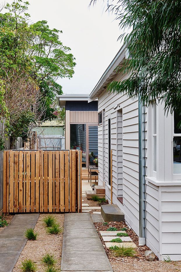 A narrow landscaped side passage between lightweight residential buildings, with a concrete footpath, stepping stones, gravel and ornamental grasses. The foreground building has pale horizontal weatherboard cladding, white trim, a pitched roof edge and a downpipe. A warm timber slatted privacy fence and gate separate the path from the garden, while a darker-clad extension and glazed doorway are visible beyond. Mature vegetation frames the passage.